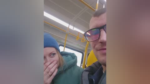 Video of a young woman in a blue beanie, green jacket, and sunglasses, and a man in glasses, sitting on a yellow bus. Both have their hands over their mouths, laughing.