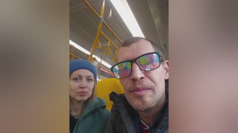 Video of a man with a shaved head and glasses, wearing a green jacket, and a woman with a blue hat, riding a yellow subway car.
