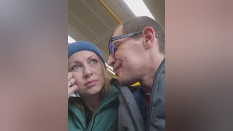Video of a blonde woman in a blue hat and green jacket, and a bald man with glasses, sitting close on a bus, both looking thoughtful.