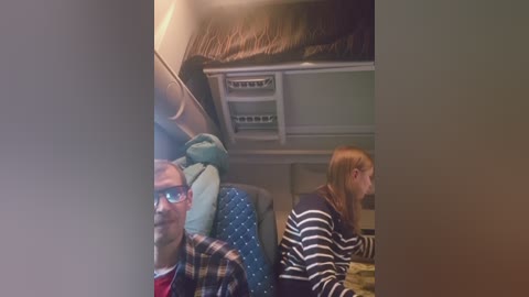 A video of two people, a man with glasses and a woman with straight hair, sitting in an airplane. They are wearing casual clothes. The background shows the cabin interior with overhead bins.