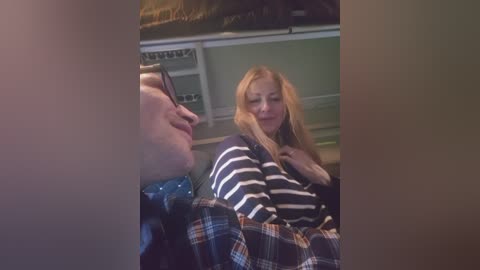 Video of a smiling woman with long blonde hair, wearing a black and white striped sweater, sitting next to a man in a plaid shirt, both inside a dimly lit, green-walled room.