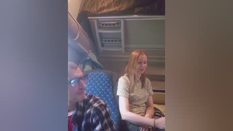 A video captures a woman with long red hair in a light yellow top, and a man in glasses and a plaid shirt, sitting in a blue cushioned seat on a bus.