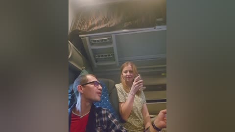 Video of a young man with glasses and short hair in a red shirt, and a woman with blonde hair in a checkered shirt, seated in a van.