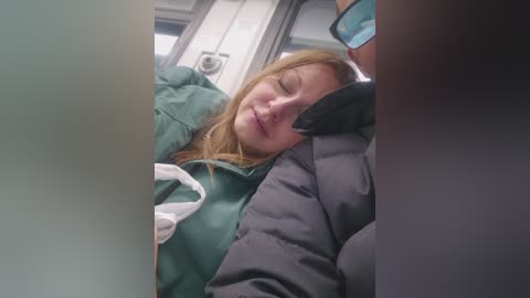 Video of a Caucasian woman with long blonde hair, wearing a green jacket and a white surgical mask, sleeping on a plane. Her head rests on a grey jacketed person's lap, with a black-gloved hand visible. The plane's interior background is visible.