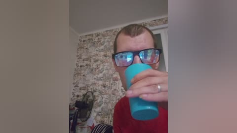 A video of a man with short hair, wearing black-framed glasses and a red shirt, drinking from a turquoise cup, in a room with a textured, patterned wallpaper background.