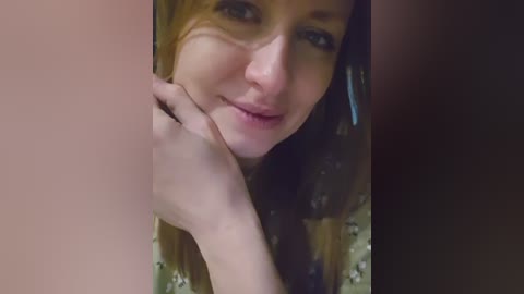 Video of a close-up of a smiling woman with light skin, blue eyes, and brown hair, partially covered by her hand, in a dimly lit room.