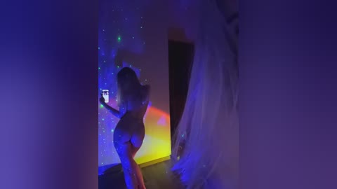 A video of a curvy, nude woman with long hair, captured from behind, standing in a dimly lit room with glowing, colorful LED lights on the wall.