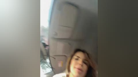 A video of a woman with light skin and brown hair, leaning against a reflective surface, with a blurred background showing a white wall and a mirror.