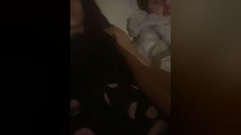 A dimly lit video captures a person in black clothing lying on a bed with a white pillow, partially covered by a white blanket.