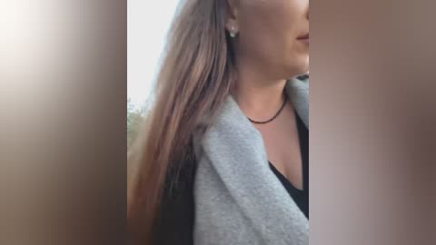 A close-up video of a woman with long, straight brown hair, wearing a gray cardigan over a black top. The background is blurred, showing an outdoor setting with soft lighting.