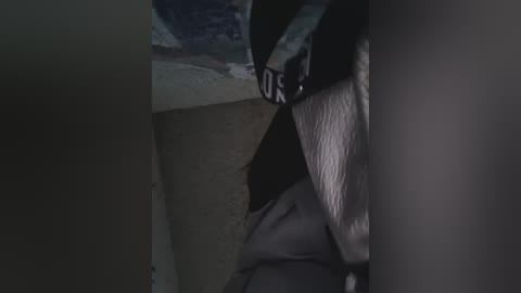 A dimly lit video of a person, possibly male, with a dark beard, wearing a gray hoodie, sitting on a concrete floor, with a black backpack beside them.