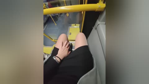 A video shows a person wearing a black dress, seated on a yellow bus seat, with a hand on their knee. The background features a bus interior with yellow railings and blue flooring.
