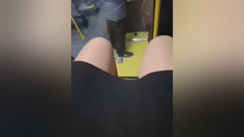 Video of a person's legs in black ribbed shorts, sitting on a yellow plastic chair inside a dimly lit room, blurred background with a hint of a person's torso in grey clothing.