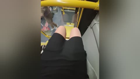 Video of a woman's legs in black shorts and knee-high socks, seated on a yellow bus seat. A reflection of a person in pink clothing is visible on the window.