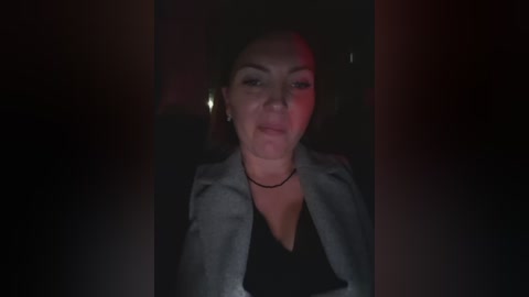 A dimly lit video of a young woman with light skin, dark hair, and a serious expression, wearing a black top and a gray blazer, standing indoors against a dark background with red lighting.