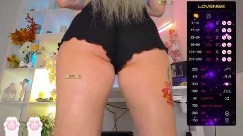 Video of a person's lower body in black shorts, with visible tattoos on their thighs, in a colorful, cluttered room.