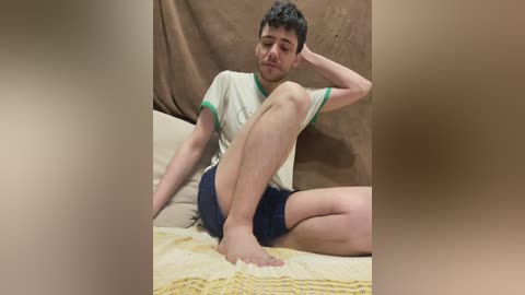 Video of a young man with short curly hair, wearing a white T-shirt and dark shorts, sitting on a bed with yellow and white blankets, against a brown fabric backdrop.