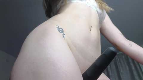 Video of a nude, fair-skinned woman with a tattoo of a snake on her right hip, holding a black dildo, taken from a low angle. Background is a dimly lit room with curtains.