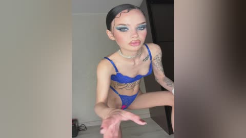 Video of a slender, fair-skinned transgender woman with dark hair, wearing blue lingerie, posing in a room. She has tattoos on her arms and chest, with makeup emphasizing her eyes.