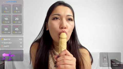 Video of a young Asian woman with long black hair, wearing a beige top, licking a large, circumcised, erect penis in a white room. Virtual fitness app interface overlays on the left.