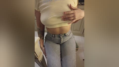 A video of a woman with fair skin, medium build, wearing a yellow crop top and light blue jeans, standing in a bathroom with tiled walls and a white cabinet.