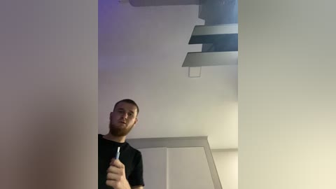 Video of a young man with short hair, beard, and glasses, wearing a black T-shirt, looking up at a modern, minimalist staircase with clean lines and gray tones.