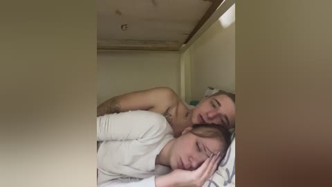 A video of a young man and woman cuddling under a blanket in a narrow, dimly lit bed, with beige walls and a wooden ceiling. The woman has blonde hair and is shirtless, while the man has glasses and a white shirt.