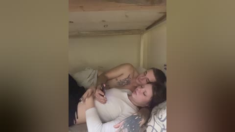 Video of a tattooed, shirtless man and a woman with long hair, both in bed, holding hands, in a dimly lit, cozy room.