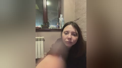 Video of a young Caucasian woman with long brown hair, wearing a black top, in a dimly lit bathroom with beige walls and a radiator.