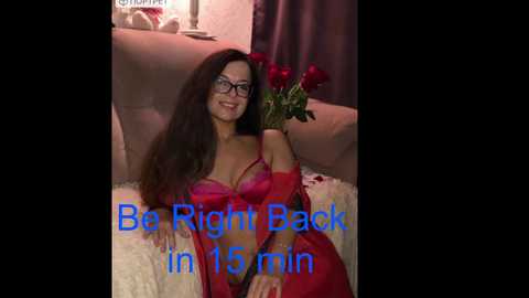 Video of a smiling woman with long brown hair, wearing glasses, red lingerie, and holding red roses, set against a pink background with a \"Be Right Back in 15 min\" text.