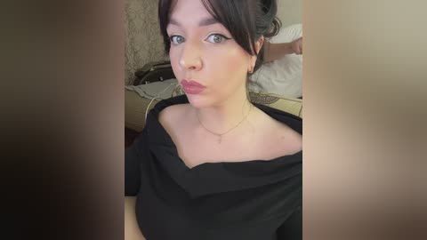 Video of a young woman with light skin and dark hair, wearing a black off-shoulder top, looking directly at the camera with a neutral expression, in a softly lit room with a bed and beige walls.