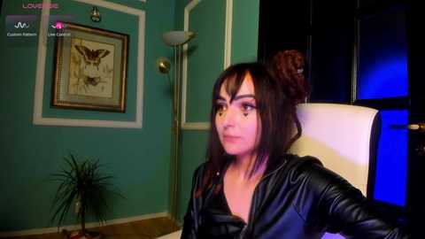 Video of a woman with dark hair in a high bun, wearing a black leather jacket, sitting in a modern room with teal walls and framed artwork, under blue lighting.