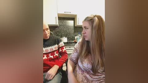 Video of an elderly man in a red sweater with white pattern, seated beside a young woman with long blonde hair, in a modern kitchen with white cabinets and granite countertops.