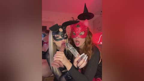 Video of two women in Halloween costumes: one in black cat ears and mask, the other in red devil mask, licking a lollipop. Background shows a dimly lit room with red lighting.