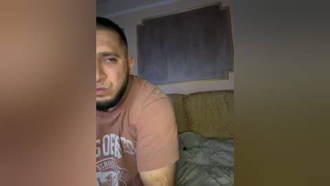 Video of a bearded man with a muscular build, wearing a maroon T-shirt, sitting on a bed with disheveled sheets, in a dimly lit room.