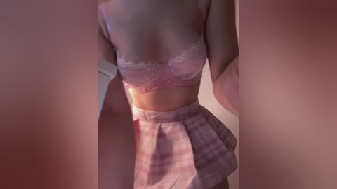 A video of a light-skinned woman in a pink plaid skirt and a lacy pink bra, standing against a blurred pink backdrop. The image is intimate and focuses on her torso and lower body.