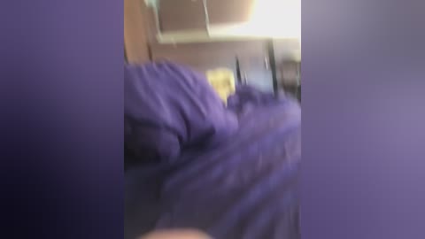 A blurry video captures a person lying on a bed with purple sheets, partially obscured by a purple wall. The background features a blurred, beige ceiling and a faint, indistinct window. The image has a soft, out-of-focus quality.