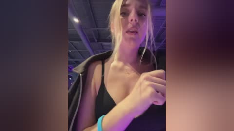 Video of a blonde woman with fair skin, wearing a black bra and jacket, holding a phone. She has a slim physique with medium-sized breasts. Background shows an industrial ceiling.