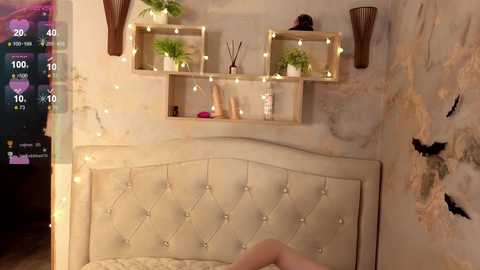 Video of a cozy bedroom with a tufted beige headboard, a wooden floating shelf adorned with plants and decor, and a textured, light-colored wall with a watercolor painting.