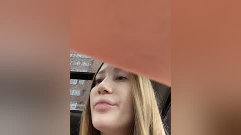 A video captures a young woman with long blonde hair, light skin, and a subtle smile, seen from the chest up. The background features a window with a brick wall outside.