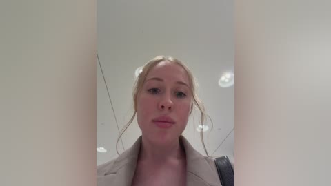 Video of a young Caucasian woman with light blonde hair styled in two buns, wearing a beige blazer, standing in a modern room with white walls and recessed lighting.