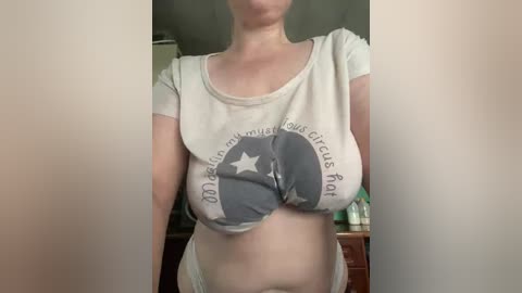 Video of a light-skinned woman with large breasts, wearing a white crop top with a circular design, and white panties, standing indoors with blurred background.