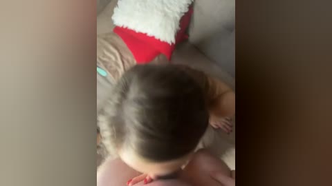 A video shows a young child with light brown hair, wearing a red top, lying on a beige couch with a white fluffy pillow and a blue toy nearby.