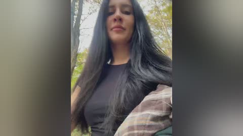 Video of a young woman with long, straight black hair, wearing a black t-shirt, standing in a forest with blurred green trees in the background.