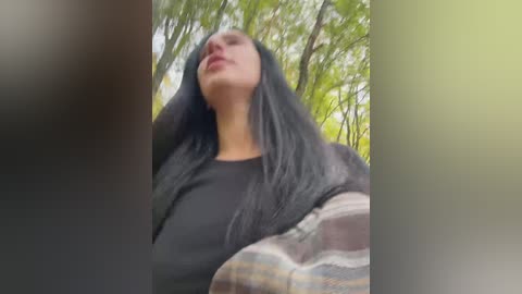 A video of a woman with long, straight black hair, wearing a black shirt, standing in a lush, green forest, with blurred trees in the background.