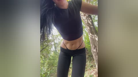 Video of a slender, fair-skinned woman with long black hair, wearing a black crop top and black leggings, standing outdoors in a forest.