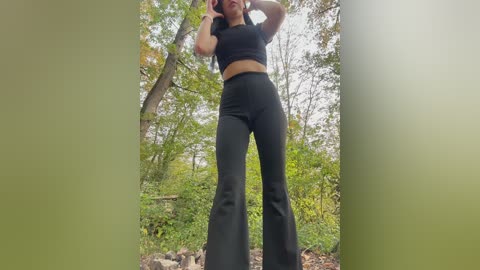 Video of a woman in black bell-bottom pants and a black crop top, posing outdoors in a lush, green forest with tall trees and sunlight filtering through the leaves.