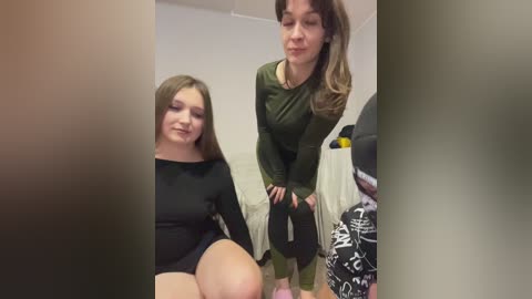 Video of two women in a bedroom. One woman in black leotard, the other in green top and pants, both with long hair.