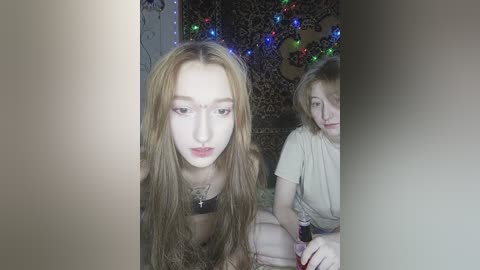 Video of two young women with long, straight hair. One wears glasses and a black top, the other a beige shirt, both indoors with a patterned wall and multicolored lights.