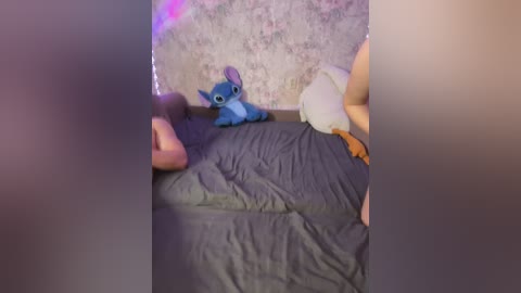Video of a bedroom with a person lying on a bed, partially covered by a gray sheet. A stuffed Stitch toy is positioned between them, and the room has floral wallpaper and a faint purple LED strip lighting.